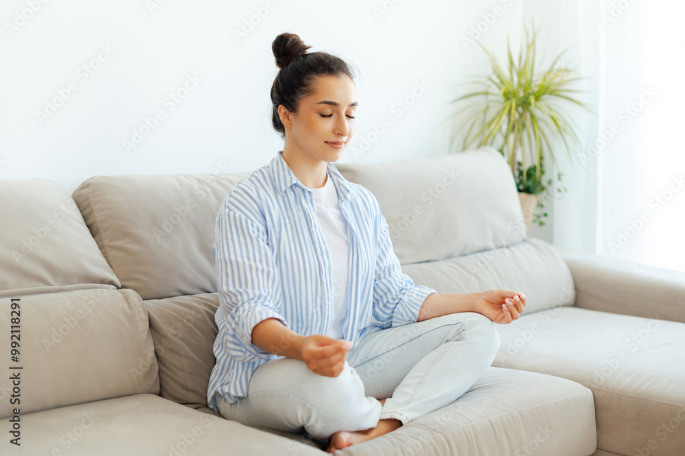 Relaxation in the morning. Calm pretty young arabian or indian woman in a cozy living room, sitting on sofa and meditating in a lotus position with eyes closed, doing yoga mindfulness training Relaxation in the morning. Calm pretty young arabian or indian woman in a cozy living room, sitting on sofa and meditating in a lotus position with eyes closed, doing yoga mindfulness training
