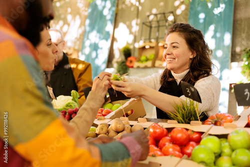 Friendly farmer offering samples to customers, showcasing homegrown fruits and vegetables at local farmers market. Young multiracial couple or family tasting natural fresh produce, visiting food fair.