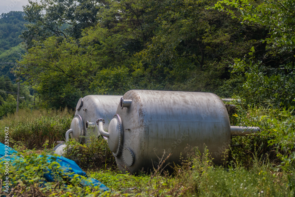 Large metal industrial tanks sit in an overgrown area, surrounded by forest greenery. Large metal industrial tanks sit in an overgrown area, surrounded by forest greenery.