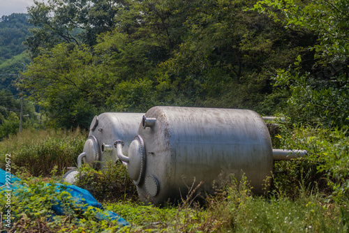Large metal industrial tanks sit in an overgrown area, surrounded by forest greenery.