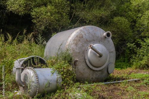 Large metal industrial tanks sit in an overgrown area, surrounded by forest greenery.