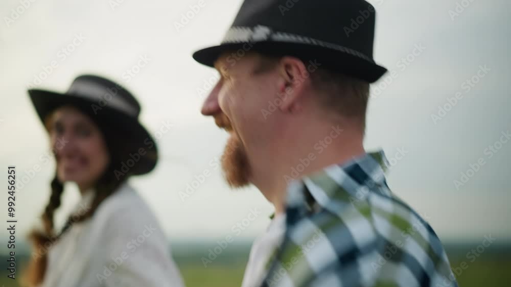 Close-up of a joyful couple running through a grassy field at sunset, smiling . The man is styled in a black hat and checkered shirt, and the woman in a white dress with a matching hat