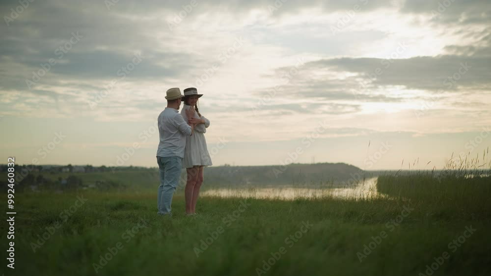 A tender moment between two sweethearts, as a man in a hat, white shirt, and jeans holds a woman in a white dress on a grassy hill at sunset. The scene exudes warmth, love, and connection A tender moment between two sweethearts, as a man in a hat, white shirt, and jeans holds a woman in a white dress on a grassy hill at sunset. The scene exudes warmth, love, and connection