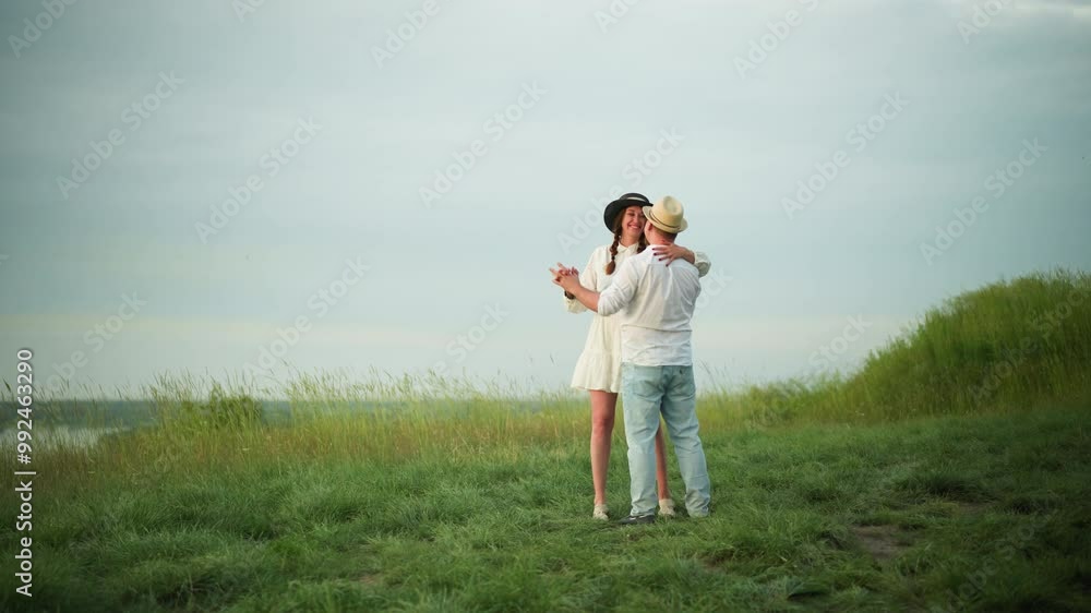 A loving couple dances together on a grassy hill, with the man wearing a white shirt, hat, and jeans, and the woman dressed in a white dress and black hat.The scene captures a tender and joyful moment A loving couple dances together on a grassy hill, with the man wearing a white shirt, hat, and jeans, and the woman dressed in a white dress and black hat.The scene captures a tender and joyful moment