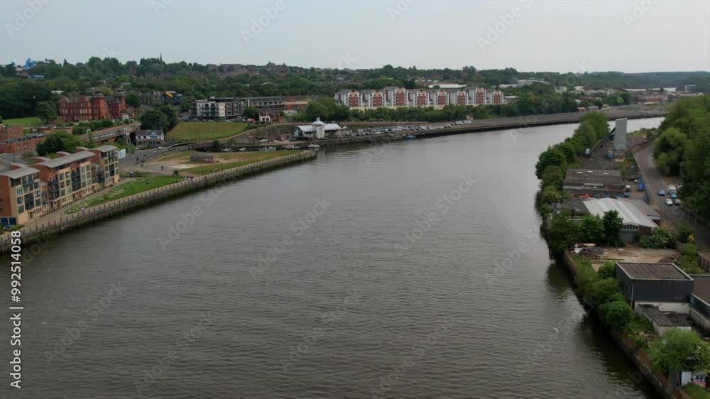 Aerial view of modern riverside apartments and an industrial area along the River Tyne, showcasing urban development and greenery. Aerial view of modern riverside apartments and an industrial area along the River Tyne, showcasing urban development and greenery.