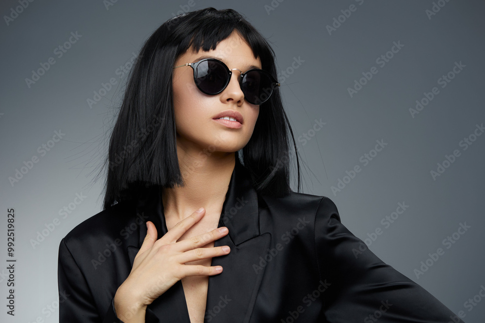 Beautiful woman in a black suit and sunglasses posing confidently against a gray background, exuding elegance and power with her stylish and sophisticated look Beautiful woman in a black suit and sunglasses posing confidently against a gray background, exuding elegance and power with her stylish and sophisticated look