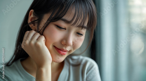 A young Asian American with a shy smile on her face and looking downwards with her eyes closed. Her cheek rests against her hand. She appears to be having a fond, pleasant memory.