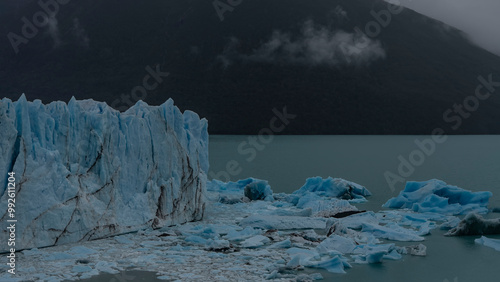 A melting glacier. A wall of cracked blue ice rises above a glacial lake. Melted ice floes float in turquoise water. The coastal mountains are in a fog. Cloudy. Perito Moreno. El Calafate. Argentina.  photo