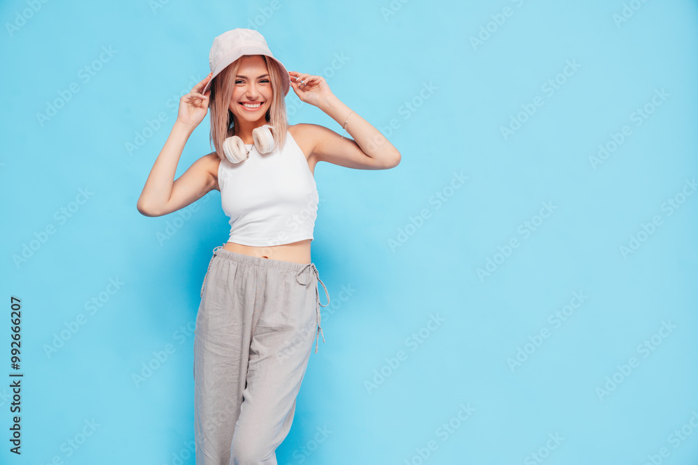 Portrait of young extremely happy blond woman wearing hipster 90s clothes and pink panama. Female standing with headphones over neck. Model isolated over blue background. Cheerful and happy, smiling Portrait of young extremely happy blond woman wearing hipster 90s clothes and pink panama. Female standing with headphones over neck. Model isolated over blue background. Cheerful and happy, smiling