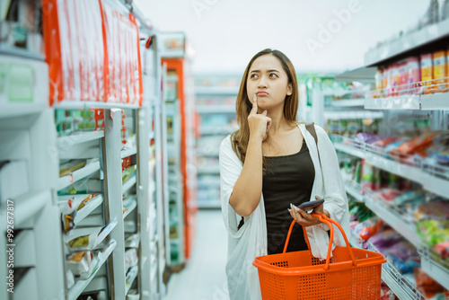 young woman thinking about what to buy while shopping carrying a cart at a minimarket photo