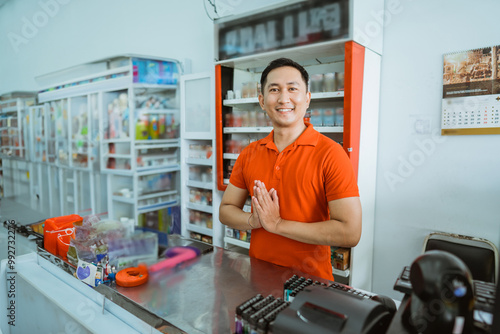 smiling male store clerk with a greeting gesture while standing at a minimarket cashier photo