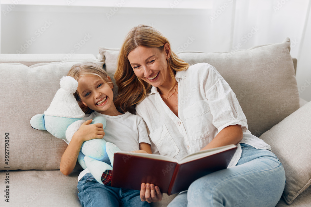 Mother and daughter bonding on the couch reading a book together while holding a teddy bear Mother and daughter bonding on the couch reading a book together while holding a teddy bear