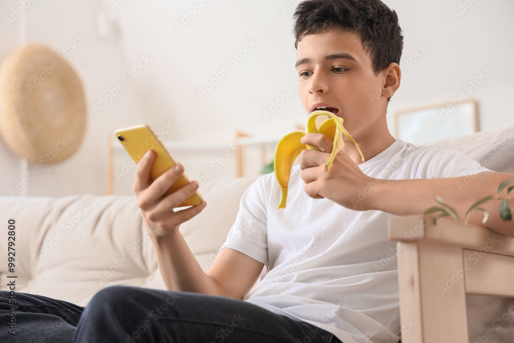 Teenage boy with mobile phone eating banana on sofa at home