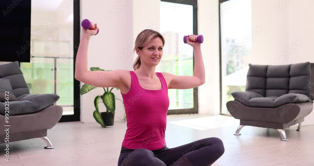 Sportive female person raises hands with dumbbells sitting on mat in modern premise of apartment. Strengthening body and maintaining health at home slow motion Sportive female person raises hands with dumbbells sitting on mat in modern premise of apartment. Strengthening body and maintaining health at home slow motion