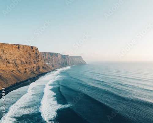 Aerial view of a dramatic coastline with rocky cliffs and crashing waves. The clear blue sky and ocean create a sense of tranquility and vastness.