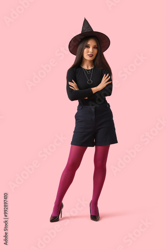 Beautiful young happy Asian woman dressed as witch for Halloween on pink background