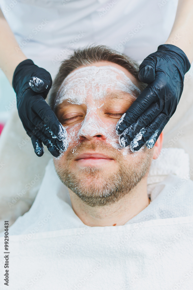 Naklejka premium A beautician applies a medical mask to a man's face. Facial skin care for men. Male cosmetology.