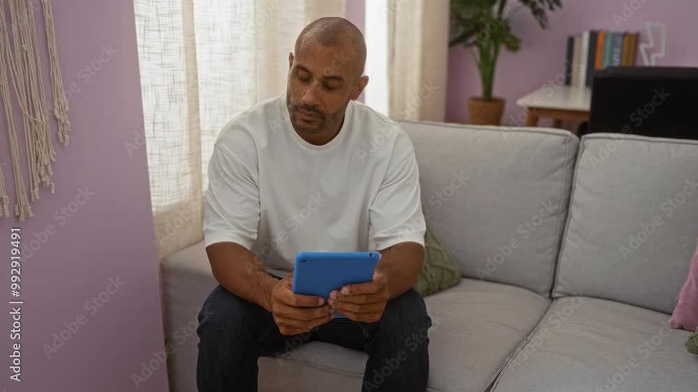 Handsome man sitting on a couch in a living room reading a tablet against a stylish decor with shelves and plants Handsome man sitting on a couch in a living room reading a tablet against a stylish decor with shelves and plants