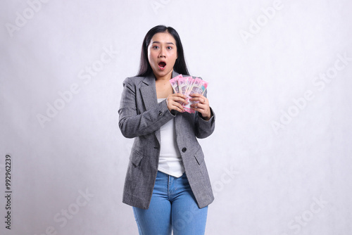 portrait of beautiful young office lady asia shocked both hands holding rupiah money wearing gray suit isolated on white background. for giveaway content, lifestyle, gift, event