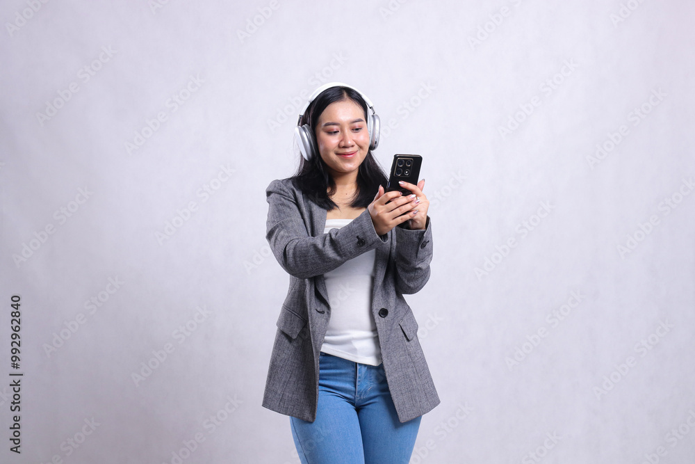 beautiful young office girl smile to gadget wearing headphone headset and holding mobile phone streaming video wearing suit isolated on white background. lifestyle concept, entertainment, technology beautiful young office girl smile to gadget wearing headphone headset and holding mobile phone streaming video wearing suit isolated on white background. lifestyle concept, entertainment, technology