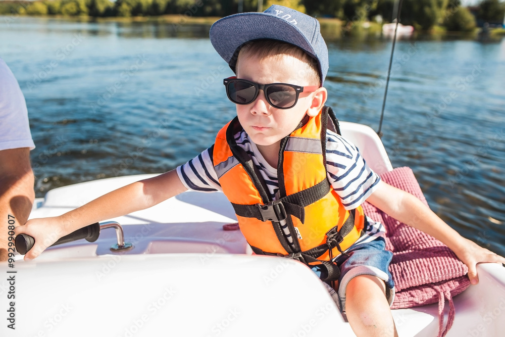 A handsome, fashionable little boy of 5 years old in a bright life jacket and sunglasses drives a catamaran on the lake. Recreation and tourism. A handsome, fashionable little boy of 5 years old in a bright life jacket and sunglasses drives a catamaran on the lake. Recreation and tourism.
