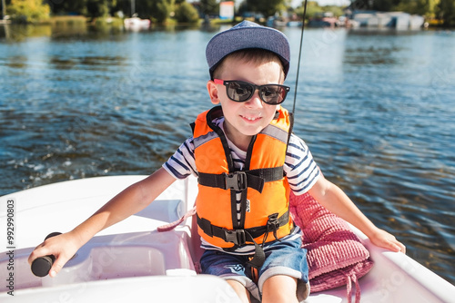 A handsome, fashionable little boy of 5 years old in a bright life jacket and sunglasses drives a catamaran on the lake. Recreation and tourism. photo