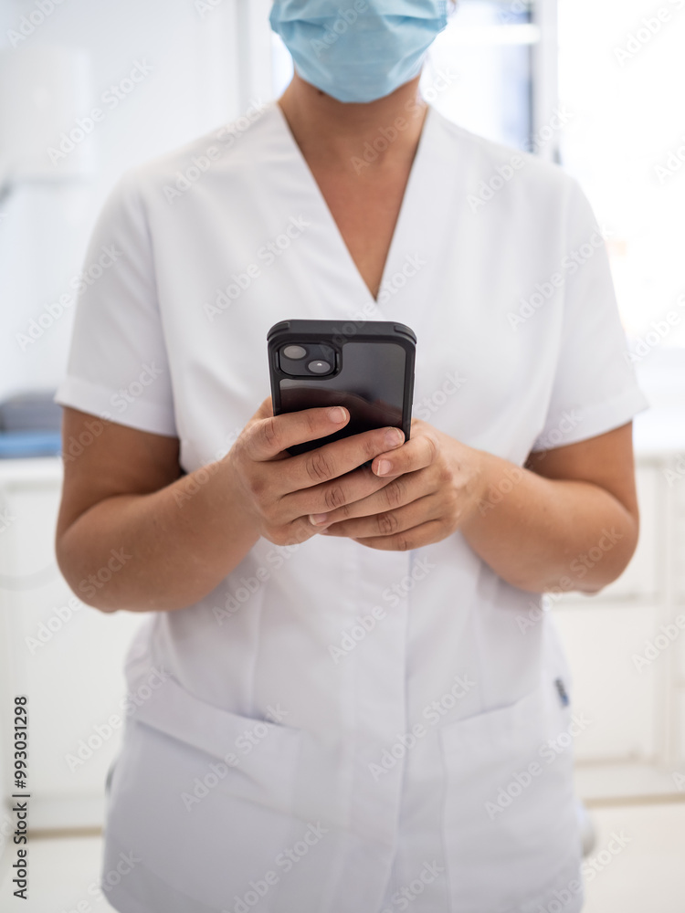 An unrecognizable healthcare professional in a white uniform and mask checks the smartphone while working in a modern clinic. The focus is on the mobile device usage. An unrecognizable healthcare professional in a white uniform and mask checks the smartphone while working in a modern clinic. The focus is on the mobile device usage.