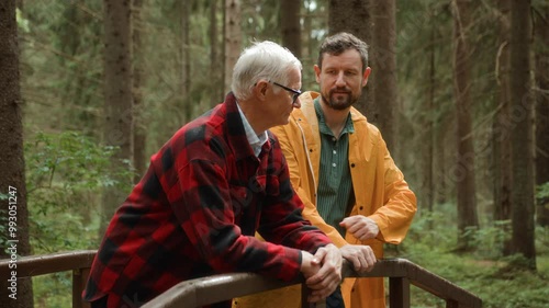 Adult son and elderly father walk together on pathway in forest, enjoying talking. Adult man supports senior people chatting and strolling. Intergenerational relationships and care for older people.