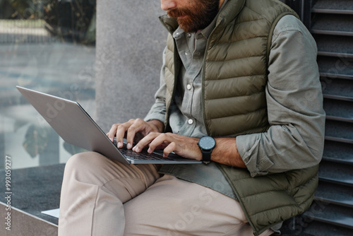 Bearded man sitting on stone bench outside, hands on laptop keyboard, wearing green vest and casual attire, focused on screen in urban setting