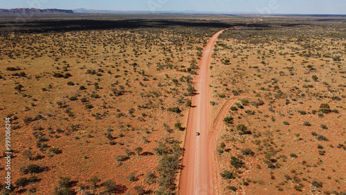Car driving on a straight gravel road in the Australian red centre. Long sandy road with trees on the side. Drone view of outback australia. Lost in the middle of the desert.