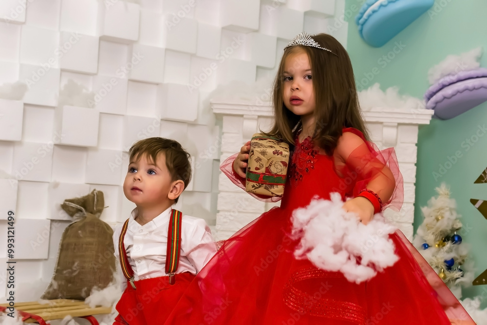 A little girl and a boy in red clothes have fun in the artificial snow by the fireplace in the studio. The girl is holding a box with gifts. Little kids in red christmas costume having fun beside a A little girl and a boy in red clothes have fun in the artificial snow by the fireplace in the studio. The girl is holding a box with gifts. Little kids in red christmas costume having fun beside a