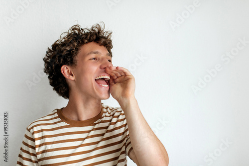 A cheerful young man expresses excitement by calling out, wearing a casual striped shirt, with a bright smile and lively energy in a simple indoor setting.