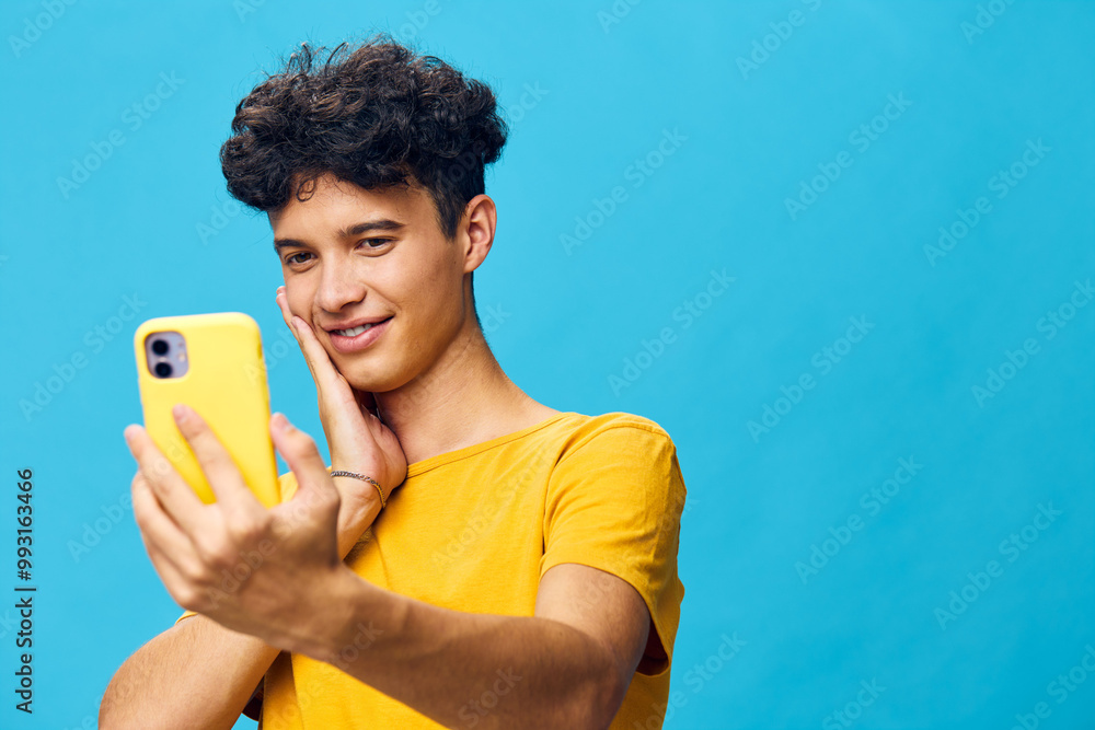 Portrait of a young man with curly hair smiling while taking a selfie with a yellow phone, set against a vibrant blue background Happy and carefree expression conveys joy Portrait of a young man with curly hair smiling while taking a selfie with a yellow phone, set against a vibrant blue background Happy and carefree expression conveys joy