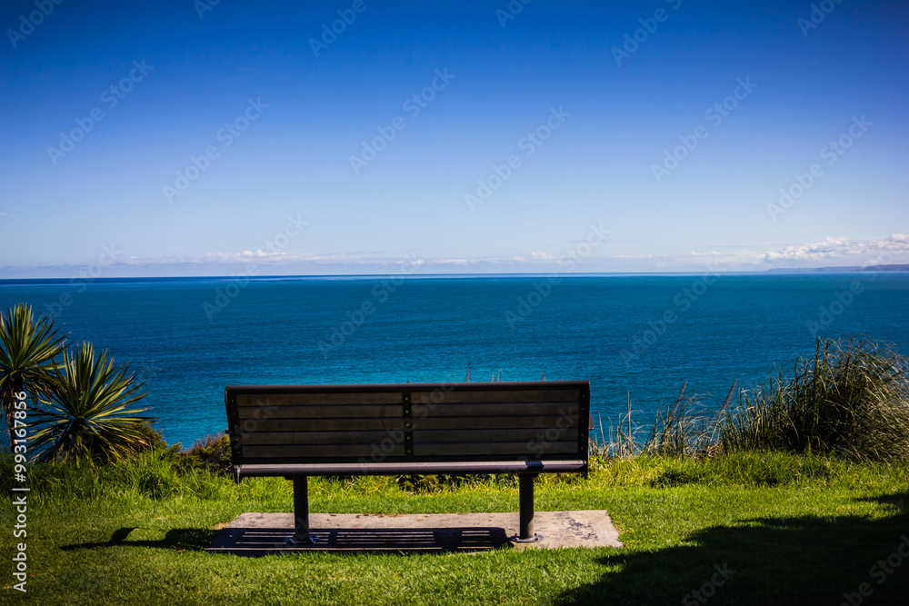 A minimalistic landscape with a bench on green grass looking out toward the ocean sea waters. Blue sky and low clouds over distant horizon. Raglan, New Zealand A minimalistic landscape with a bench on green grass looking out toward the ocean sea waters. Blue sky and low clouds over distant horizon. Raglan, New Zealand