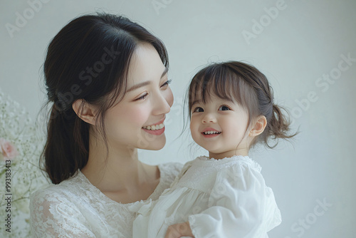 Young Chinese mother and cute daughter interacting and smiling in front of a plain background, dressed in simple Uniqlo clothes, during a family photo portrait shoot.