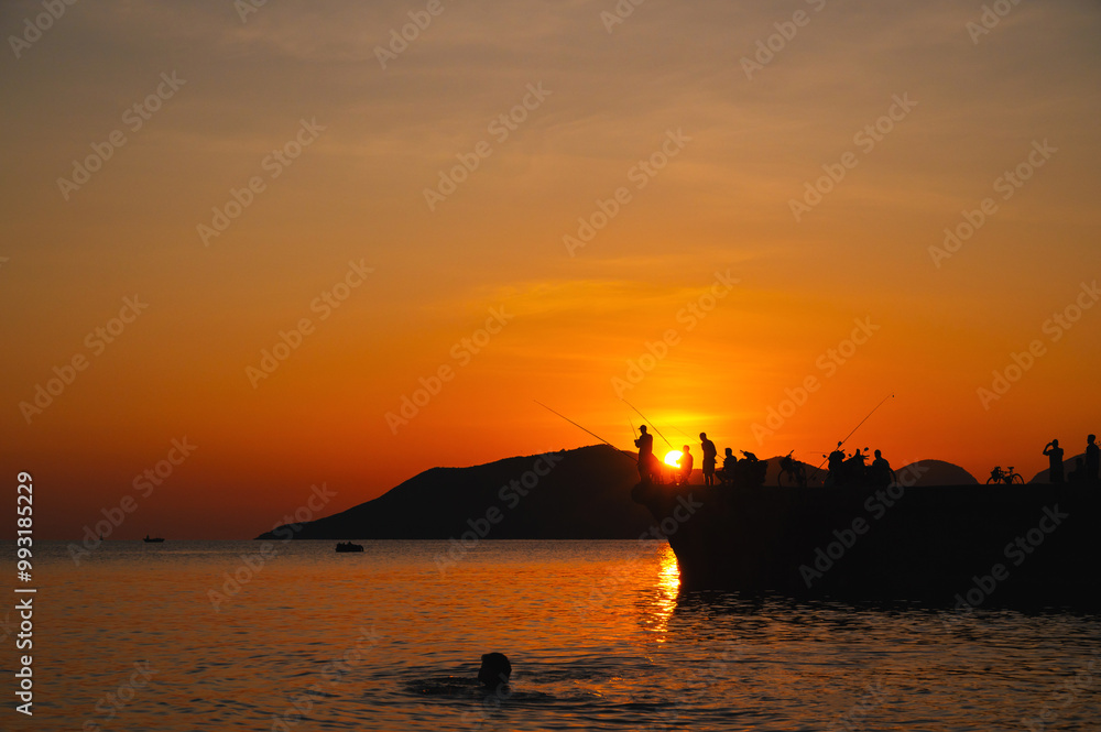 Silhouettes of fishermen with fishing rods fishing on pier in sea in the evening at sunset in summer Silhouettes of fishermen with fishing rods fishing on pier in sea in the evening at sunset in summer