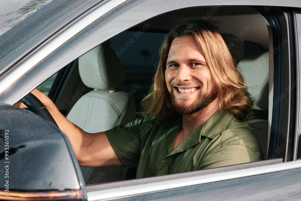 Smiling man with long hair sitting in a car, looking cheerful and confident The interior features a light color scheme Smiling man with long hair sitting in a car, looking cheerful and confident The interior features a light color scheme