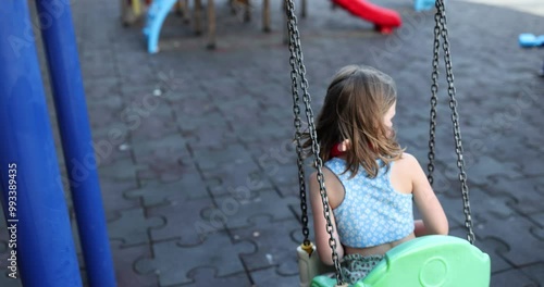 Little girl rides on swing against children slide on modern playground at daytime. Concept of entertainment and recreation in childhood slow motion photo