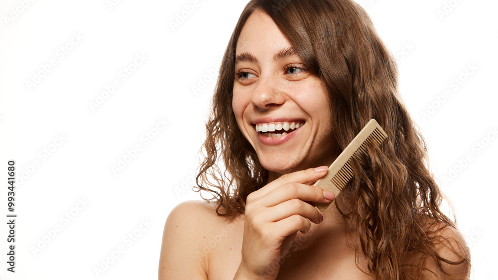 Smiling young woman combing her hair, showing joy in natural beauty and wellness moment against white studio background. Concept of beauty, fashion, appearance, self care. Ad