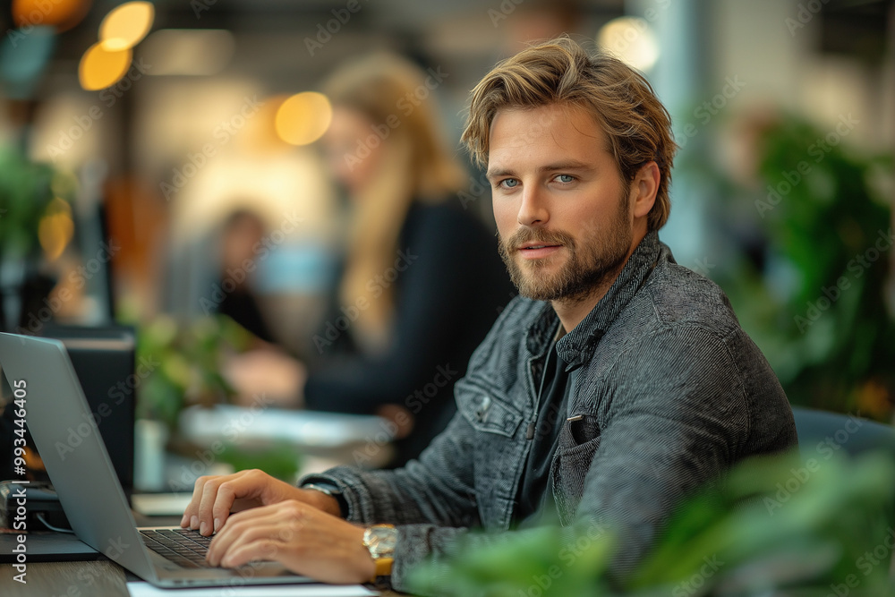 A man in a gray jacket is seated at a desk, working on a laptop. Other people are out of focus in the background A man in a gray jacket is seated at a desk, working on a laptop. Other people are out of focus in the background