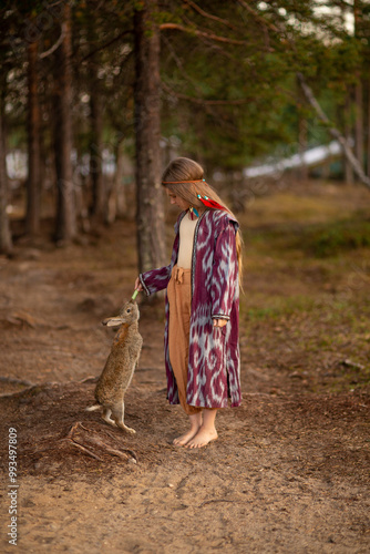 A girl with long white hair in an ethnic burgundy raincoat feeds a rabbit with a cucumber. A girl barefoot against the background of pine trees