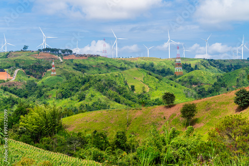 A panoramic view of a wind farm nestled amidst rolling green hills. White wind turbines stand tall against a backdrop of blue sky dotted with fluffy clouds. The scene is both serene and powerful,