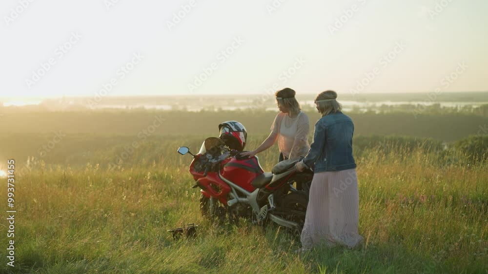 Two sisters joyfully clean a red motorcycle in an open grassy field at sunset, one sister dances while holding her gown Two sisters joyfully clean a red motorcycle in an open grassy field at sunset, one sister dances while holding her gown