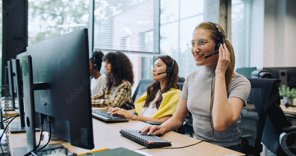 © ihorvsn - Pretty worker wearing headset during online consultation with her client. Enjoying providing quality services to customers. Beside woman sitting coworkers at their computers.