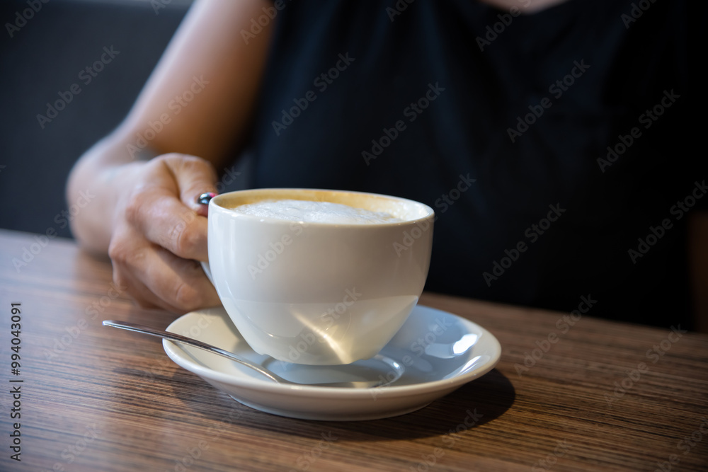Side view of female hand holding white porcelain cup of cappuccino or latte coffee drink on brown wooden table in cafe or restaurant. Soft focus. Copy space. Hot drinks theme. Side view of female hand holding white porcelain cup of cappuccino or latte coffee drink on brown wooden table in cafe or restaurant. Soft focus. Copy space. Hot drinks theme.