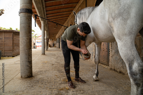 Stable hand cleaning white horseshoes in the vicinity of a stable on a sunny day.