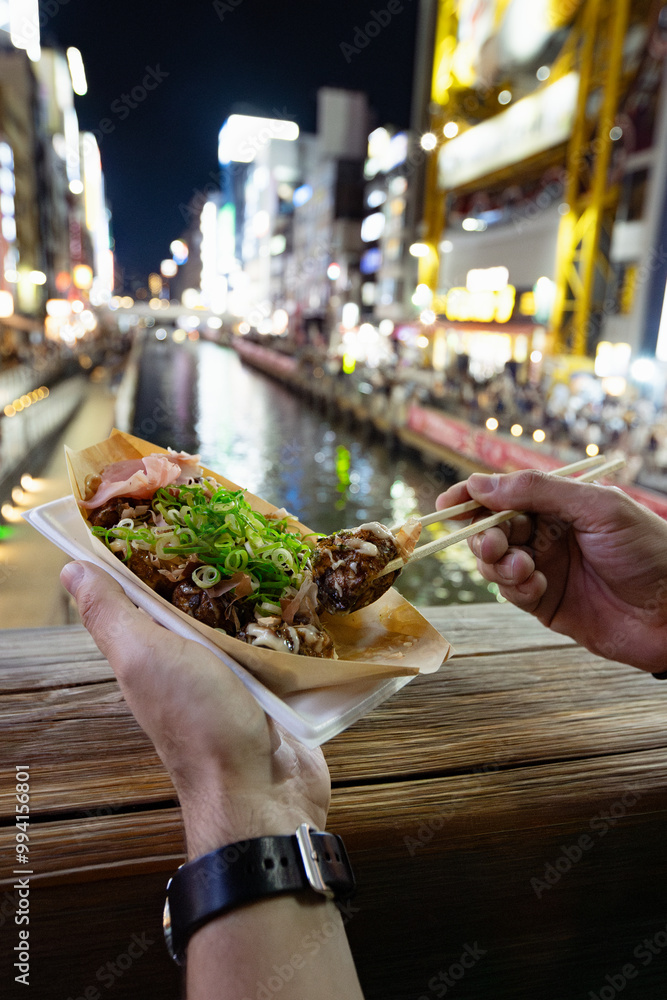 A tourist is eating a takoyaki ball with chopsticks, popular street food at Dotonbori canal in Osaka, Japan. It's a summer night, the lights of billboards are reflexing at the water in the background