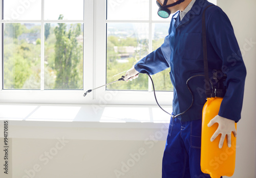 Male exterminator from pest control service wearing protective safety mask and blue workwear suit working inside house spraying toxic liquid or gas from yellow sprayer bottle over white window sill photo