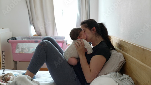 Photos Mother lying on bed while holding her baby, sharing a tender kiss with natural daylight through window in the background