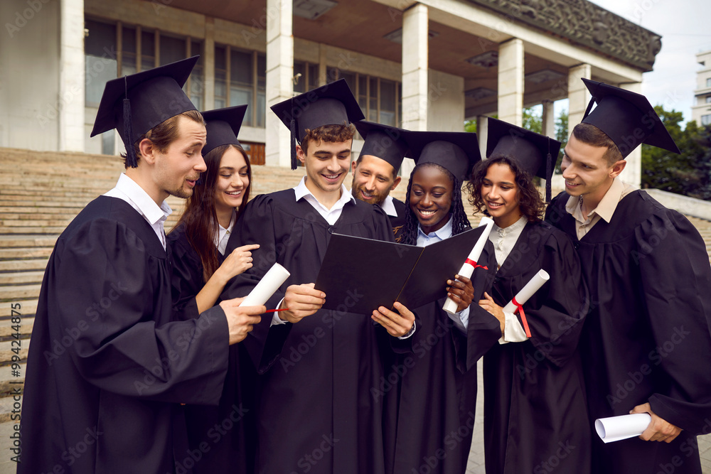 Portrait of a happy diverse multiethnic graduate students in black graduation robes celebrate graduation and looking to diploma of their classmate in university campus standing outdoors. Portrait of a happy diverse multiethnic graduate students in black graduation robes celebrate graduation and looking to diploma of their classmate in university campus standing outdoors.
